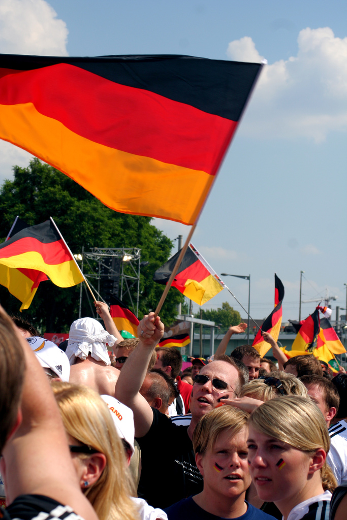 Auf dem Bild sind mehrere Fans. Sie jubeln für die deutsche Fussballmannschaft. Manche haben die Deutschlandflagge dabei. Vorne auf dem Bild sind zwei Fans die sich die Deutschlandflagge auf die Backe gemalt haben.