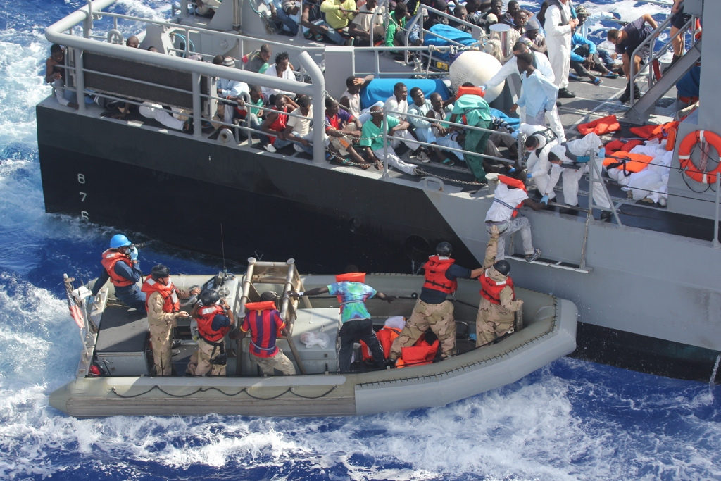Bild Nummer 1. Auf dem Bild sind Flüchtlinge und Helfer auf einem Boot und einem Schiff zu sehen
