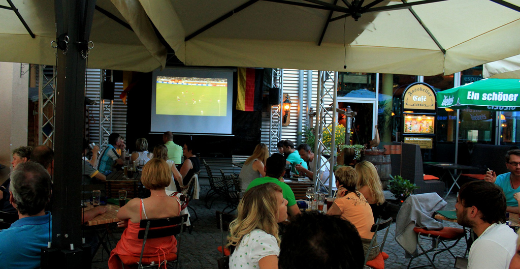 Auf dem Bild sind mehrere Personen in einer Gaststätte zu sehen. Im Fernseher läuft Fußball.